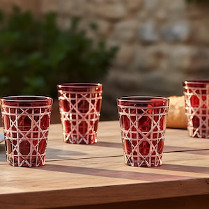 Könnte beinhalten: Vier rote Trinkgläser mit Gittermuster stehen auf einem Holztisch. Die Gläser sind mit einer dunklen Flüssigkeit gefüllt. Auf dem Tisch befinden sich auch geschnittenes Brot, Feigen und Käse, was auf ein ungezwungenes Treffen im Freien hindeutet.