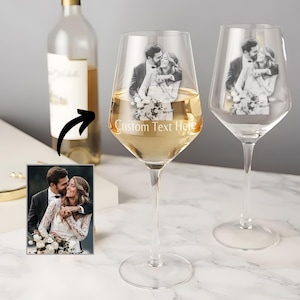 May include: Two clear wine glasses with a photo of a couple etched on the glass. One glass is filled with a light yellow liquid. The words "Custom Text Here" are printed on the glass. A bottle of wine and a photo of the couple are in the background.