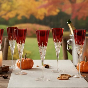 May include: A table set for a celebration, featuring six champagne flutes with ruby red bowls and a clear, patterned stem. Pumpkins, candles, and a champagne bottle in a silver ice bucket create an autumnal atmosphere. The scene is set outdoors.
