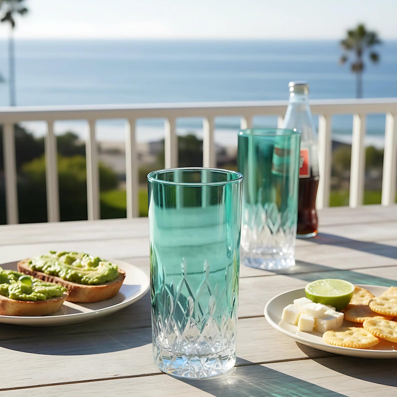 Puede incluir: Dos vasos altos de color turquesa con patrones de corte de diamante sobre una mesa de madera. Tambi&eacute;n hay una botella de refresco y platos de comida. El fondo muestra una barandilla blanca y el mar.