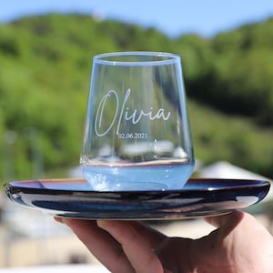 Op de afbeelding: Een helder glas met de naam "Olivia" en de datum "02.06.2021" erop gegraveerd. Het glas staat op een blauwe en bruine keramische schotel.