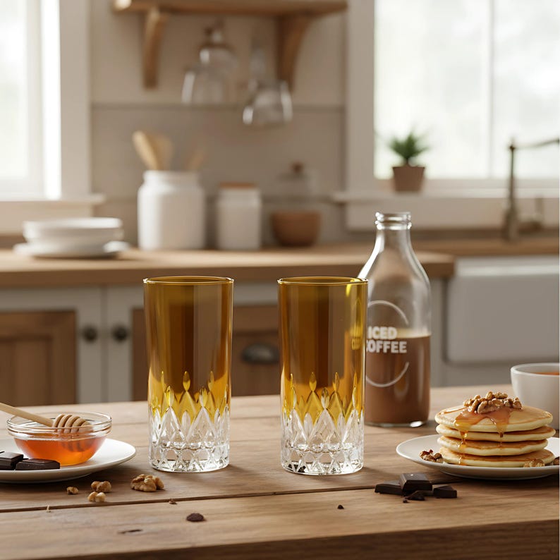 Puede incluir: Dos vasos altos y elegantes con la parte superior dorada y la base transparente tallada en diamante est&aacute;n sobre una mesa de madera. Una botella de caf&eacute; helado, panqueques y un cuenco de miel tambi&eacute;n est&aacute;n sobre la mesa, creando una escena de desayuno.
