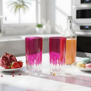 Puede incluir: Dos vasos altos de color magenta con un dise&ntilde;o tallado. Una botella transparente con l&iacute;quido naranja y un plato de macarons en el fondo. Un plato de fresas y una granada a la izquierda.
