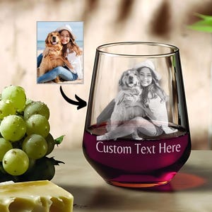 Op de afbeelding: Een helder glazen wijnglas met een gepersonaliseerde foto van een persoon en een hond gegraveerd in het glas. Het glas is gevuld met rode wijn en heeft de tekst "Custom Text Here" gegraveerd op de bodem.