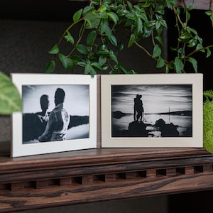 May include: Two black and white photographs in a brown frame. The left photo shows a couple standing on a beach, facing the camera. The right photo shows a couple standing on a beach, facing away from the camera, with a sunset in the background.