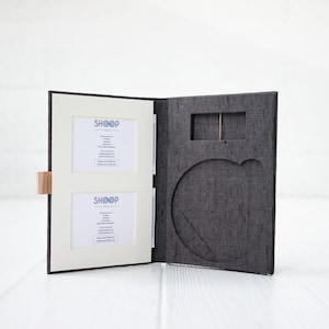 Può includere: Album fotografico aperto con due cornici e una copertina grigio scuro. L'album ha una chiusura in legno chiaro e la parola "SHOOP" è stampata sui telai bianchi. L'album è aperto su una superficie bianca.