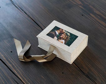 May include: A beige linen box with a brown ribbon. The box has a clear window that shows a photo of a couple embracing.