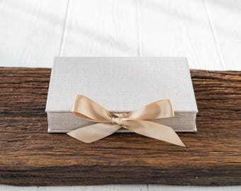 May include: A beige linen box with a brown satin ribbon tied in a bow. The box is sitting on a wooden surface.