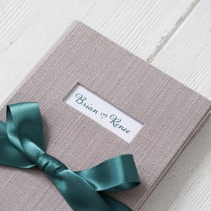 Può includere: Un album fotografico rivestito in lino grigio con un nastro turchese legato intorno. L'album ha una finestra sul davanti con il testo "Brian ♥ Renee" all'interno.