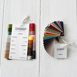 Può includere: Una cartella colori di nastri e tessuti di Shoop. La cartella dei nastri mostra 12 colori diversi di nastri, tra cui bianco, oro, marrone e viola. La cartella dei tessuti mostra 20 colori diversi di tessuti, tra cui beige, grigio, blu e rosso.