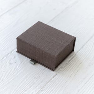 Può includere: Una piccola scatola rettangolare con una copertura in tessuto marrone. La scatola ha una linguetta di trazione argentata.