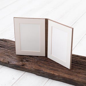 May include: Two brown picture frames with white mats and blank white inserts. The frames are open and sitting on a piece of weathered brown wood.