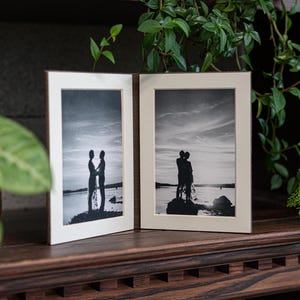 May include: Two black and white photographs in simple frames, each showing a couple silhouetted against a sunset on a beach.
