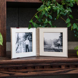 May include: A linen-covered photo album with two black and white photographs of a couple. The album is open and the photos are visible. The photos show the couple in a field and in a forest.