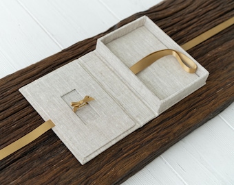 May include: A linen-covered box with a gold ribbon and a small cutout for a ring. The box is open and the ribbon is looped inside.