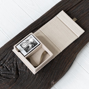 May include: A black and white photograph in a small, open, beige box. The box is inside a larger, open, beige box. The boxes are on a dark brown, textured wooden surface. The photograph shows two people.