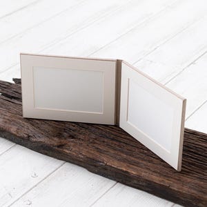 May include: Two white picture frames with a light brown border are attached together. The frames are sitting on a piece of weathered brown wood.