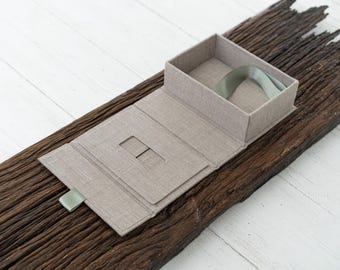 May include: A beige linen box with a green ribbon. The box is open and has a small slot in the front. The box is sitting on a piece of weathered wood.