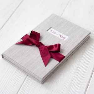 May include: A gray linen-covered photo album with a burgundy ribbon tied in a bow. The album has a window on the front with the text "Blake & Molly" printed inside.