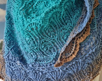 Puede incluir: Un chal hecho a mano, de tejido abierto, en tonos turquesa, azul y marrón. El chal presenta un patrón detallado y calado, con un borde festoneado. El degradado de colores añade profundidad al diseño, creando un accesorio único.