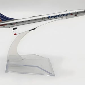 Può includere: Modello argentato di un aereo Concorde American. Il modello presenta il logo della compagnia aerea e l'iconico design della coda rosso, bianco e blu. L'aereo è montato su un supporto trasparente e curvo.