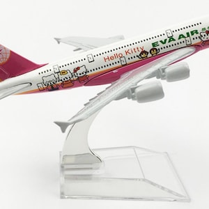 May include: A model airplane with a white fuselage and pink accents, featuring Hello Kitty characters and the text "EVA AIR." The tail fin has a pink and white design. The plane is mounted on a clear plastic stand.