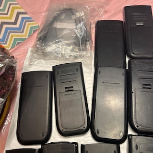 May include: A close-up of multiple black graphing calculators. The calculators have a silver trim and a small screen. The calculators are arranged in a pile on a pink surface.