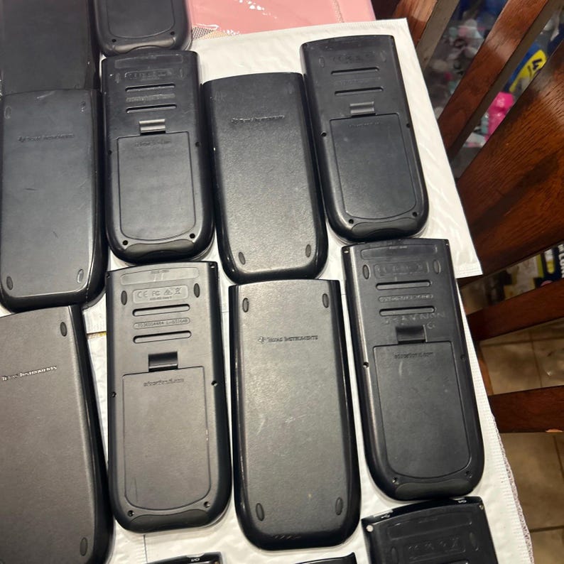 May include: A collection of ten black graphing calculators. The calculators are all in the same style and have a similar design. The calculators are arranged in a row, with the top row having five calculators and the bottom row having five calculators.