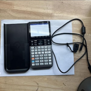 May include: A black and silver graphing calculator with a screen and a keypad. The calculator is in a black case and has a black cable attached to it.