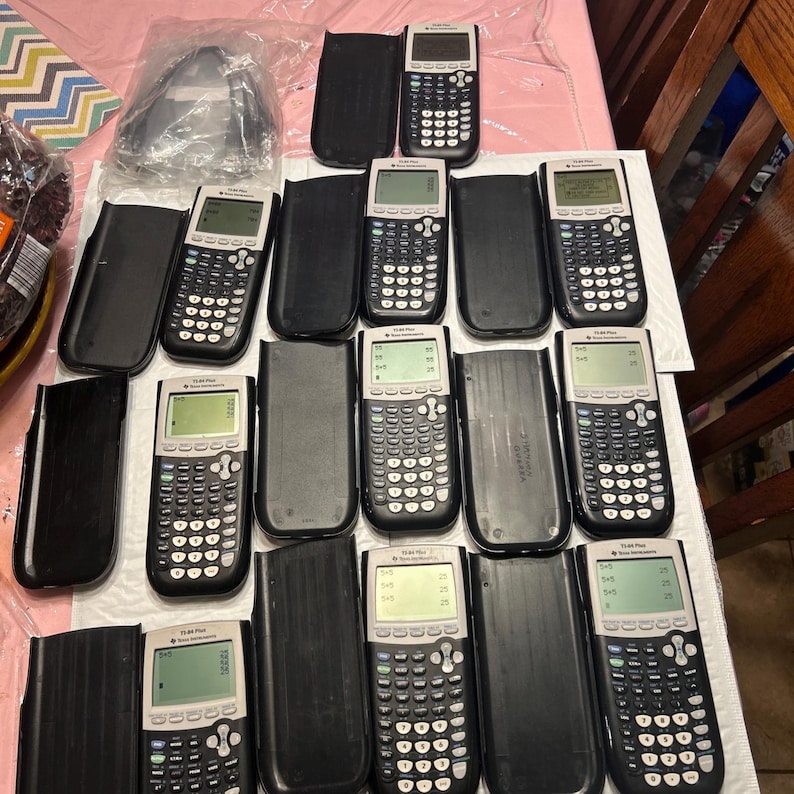 May include: A collection of 12 black and silver TI-84 Plus graphing calculators in protective cases. The calculators have a black and white screen and a variety of buttons.