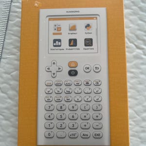 Pode incluir: Uma calculadora gráfica branca NumWorks com um ecrã digital que mostra ícones para Calculs, Grapheur, Python, Statistiques, Probabilités e Equations. O dispositivo possui um pad direcional, um botão home e um teclado completo.