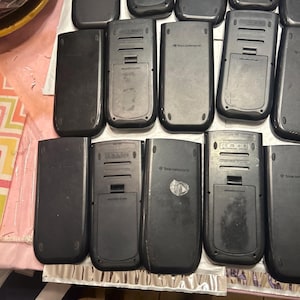 May include: A close-up of the back of several black graphing calculators. The calculators have a silver trim and the words "Texas Instruments" are printed on the back.