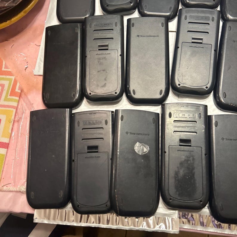 May include: A close-up of the back of several black graphing calculators. The calculators have a silver trim and the words "Texas Instruments" are printed on the back.
