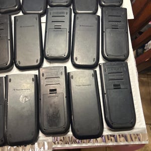 May include: A collection of ten black graphing calculators, all with the same design. The calculators have a silver screen and a black and silver keypad. The calculators are arranged in two rows of five.