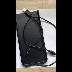 Könnte beinhalten: Ein schwarzes USB-Kabel ist in einem schwarzen rechteckigen Gehäuse mit einem USB-Stecker an einem Ende aufgewickelt.