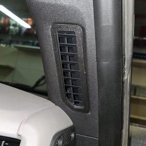 Puede incluir: Primer plano de una rejilla de ventilaci&oacute;n negra en el interior de un coche. La rejilla es rectangular con m&uacute;ltiples lamas horizontales. El embellecedor circundante es texturizado y oscuro, contrastando con los componentes interiores m&aacute;s claros.
