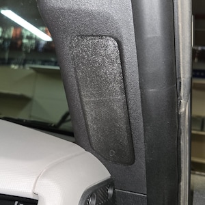 Puede incluir: Detalle interior de un coche con un panel de pl&aacute;stico negro texturizado y una cubierta rectangular para altavoz. El panel est&aacute; montado en el pilar del coche, cerca del parabrisas y el salpicadero. El salpicadero es de color gris claro.