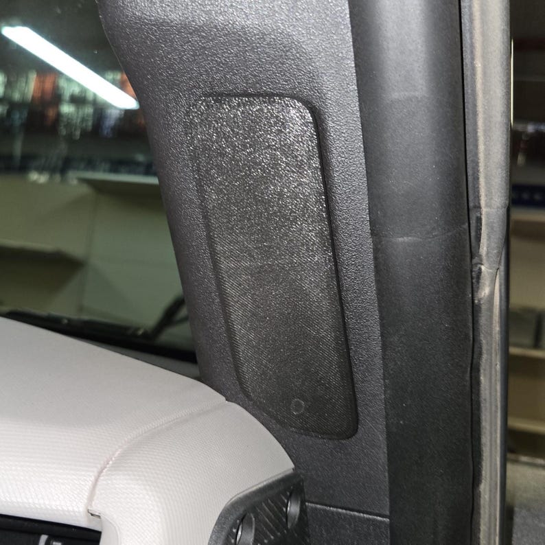 Puede incluir: Detalle interior de un coche con un panel de pl&aacute;stico negro texturizado y una cubierta rectangular para altavoz. El panel est&aacute; montado en el pilar del coche, cerca del parabrisas y el salpicadero. El salpicadero es de color gris claro.