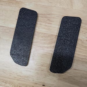 Puede incluir: Dos almohadillas rectangulares negras texturizadas con esquinas redondeadas se muestran sobre una superficie de madera de color marr&oacute;n claro. Las almohadillas tienen una forma ligeramente irregular en un extremo.