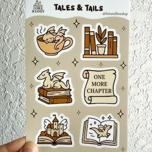 Puede incluir: Una hoja de pegatinas con el texto "TALES & TAILS" e ilustraciones de dragones, libros y un castillo. Las pegatinas son en tonos marrones y beige, con un estilo caprichoso y de dibujos animados. La hoja incluye el texto "ONE MORE CHAPTER".