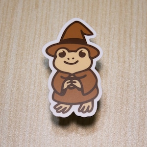 May include: A sticker featuring a cartoon frog wearing a brown wizard hat and robe. The frog has a smiling face and is holding its hands together. The sticker has a white border and is set against a light wood grain background.