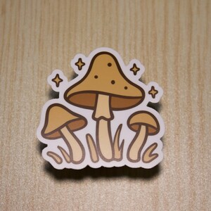 Op de afbeelding: Een sticker met drie gestileerde paddenstoelen in bruin- en beigetinten, met een witte omtrek. De grootste paddenstoel heeft een gestippelde kap en is omgeven door vier stervormige sparkles. De sticker ligt op een licht houten oppervlak.
