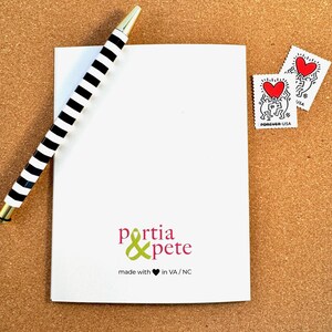 Peut inclure: Une carte blanche avec un stylo ray&eacute; noir et blanc dessus. La carte porte un logo vert et rose qui dit "portia & pete" et "made with &hearts; in VA/NC".