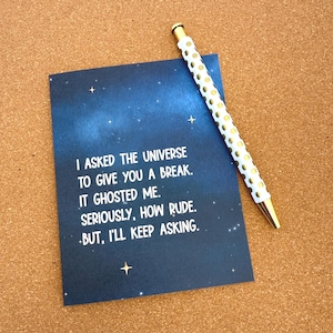 May include: A navy blue greeting card with white text that reads "I asked the universe to give you a break. It ghosted me. Seriously, how rude. But, I'll keep asking." A white pen with gold polka dots rests on the card.