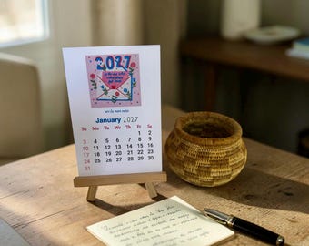 May include: A 2027 desk calendar on a small wooden easel featuring a pink illustration of an envelope with flowers and the text 2027, Be the one who makes others feel loved, and write more notes, displayed above the January 2027 calendar grid.