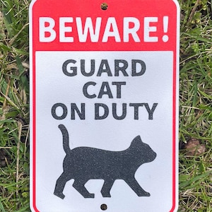 Puede incluir: Un letrero rojo y blanco con el texto "BEWARE! GUARD CAT ON DUTY" y una silueta negra de un gato caminando.