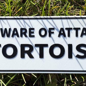 May include: A rectangular white sign with black text that reads "BEWARE OF ATTACK TORTOISE." The sign has rounded corners and four holes, with a black border. The sign is placed on a bed of green grass.