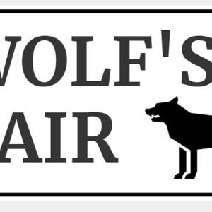 May include: White rectangular sign with black text that reads "WOLF'S LAIR". A black silhouette of a wolf is on the right side. The sign has a black border and two silver rivets. A decorative item for a home or cabin.
