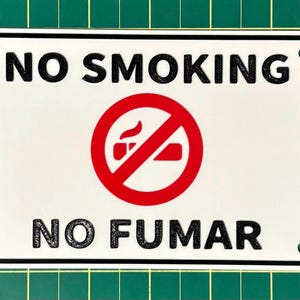Könnte beinhalten: Ein rechteckiges weißes Schild mit abgerundeten Ecken, mit den Worten "NO SMOKING" in fetten schwarzen Buchstaben über "NO FUMAR". Ein roter Kreis mit einem durchgestrichenen Zigarettensymbol befindet sich in der Mitte. Das Schild hat einen schwarzen Rand und ist mit drei Nieten befestigt.