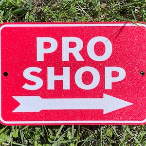 Peut inclure: Un panneau rouge avec du texte blanc et une flèche pointant vers la droite. Le panneau indique "PRO SHOP".